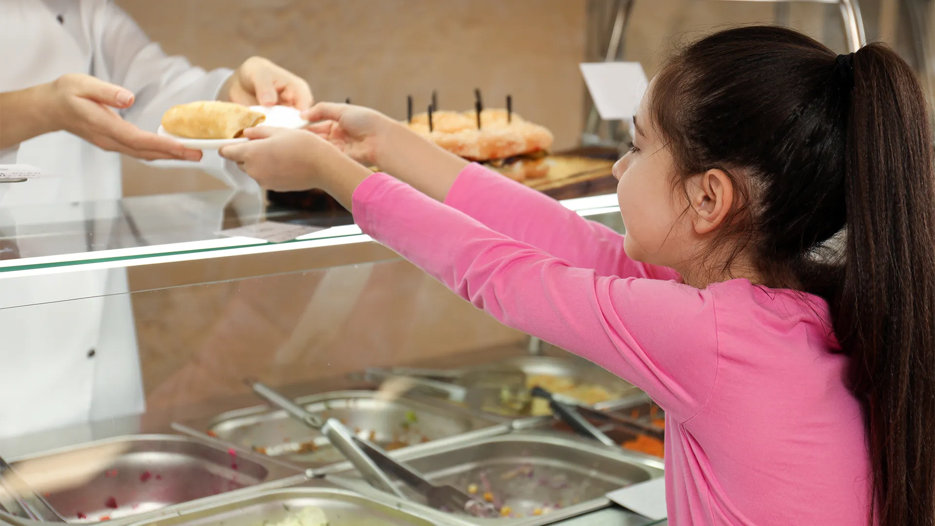 elementary school girl in pink shirt reaches for a plate with a biscuit from a school cafeteria worker
