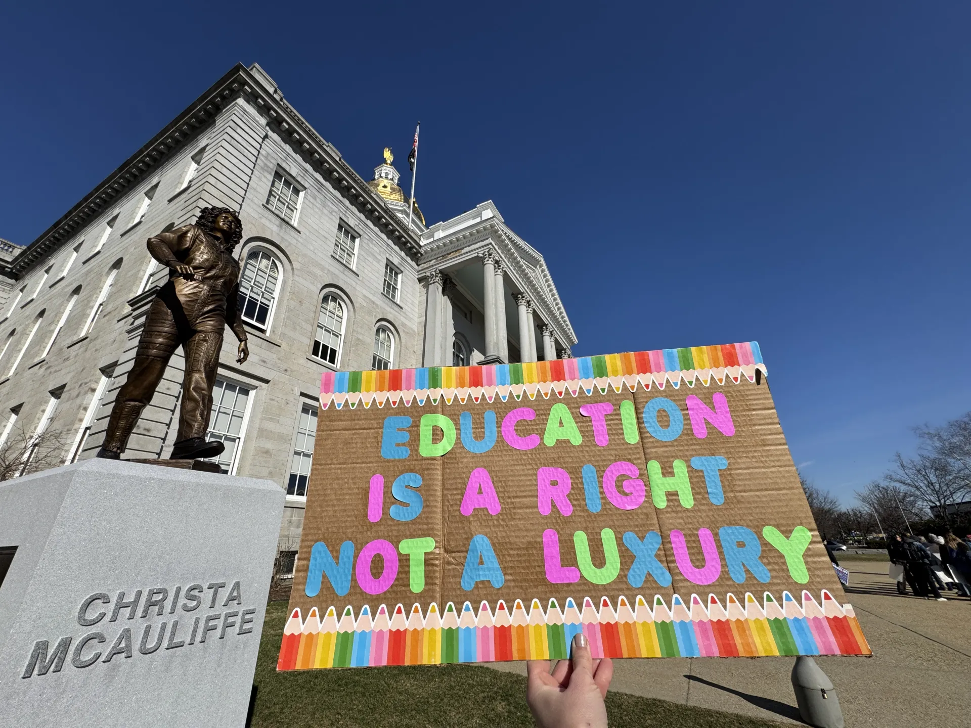 An image of a sign that reads "Education is a Right, Not a Luxury" with the State House and Christa McAuliffe statue in the background.