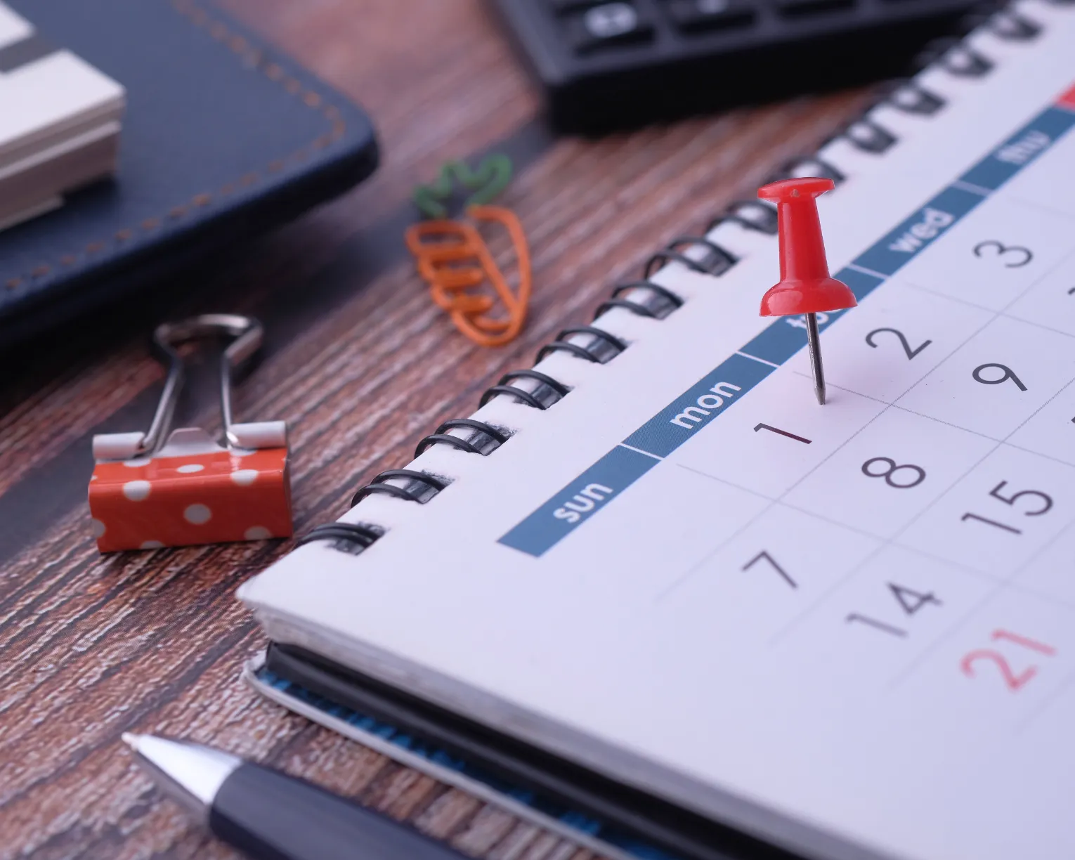 A desk calendar lays on a wood desk with a push-pin on the 2nd. Paper and binder clips are on the desk next to it.