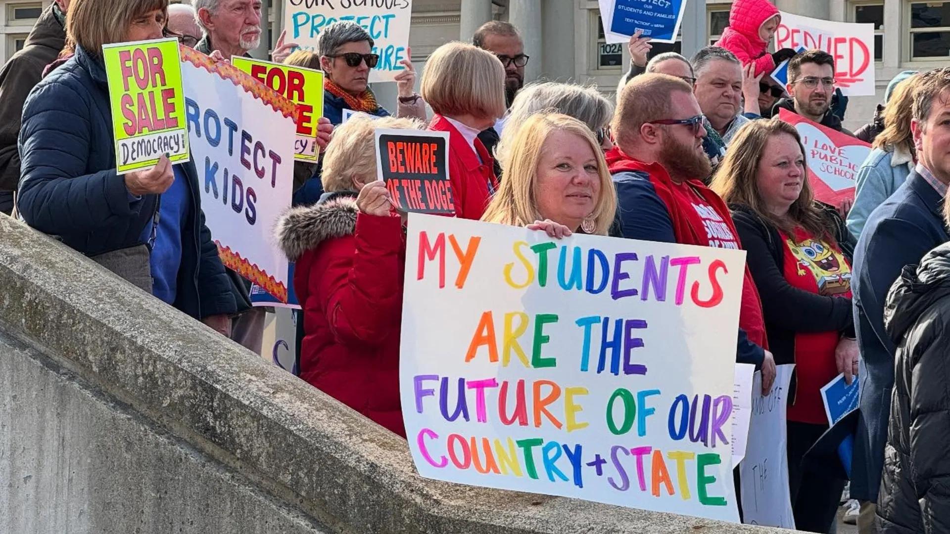 At a school walk-in on March 19, 2025, an educator holds a colorful sign that says "My students are the future of our country + state."