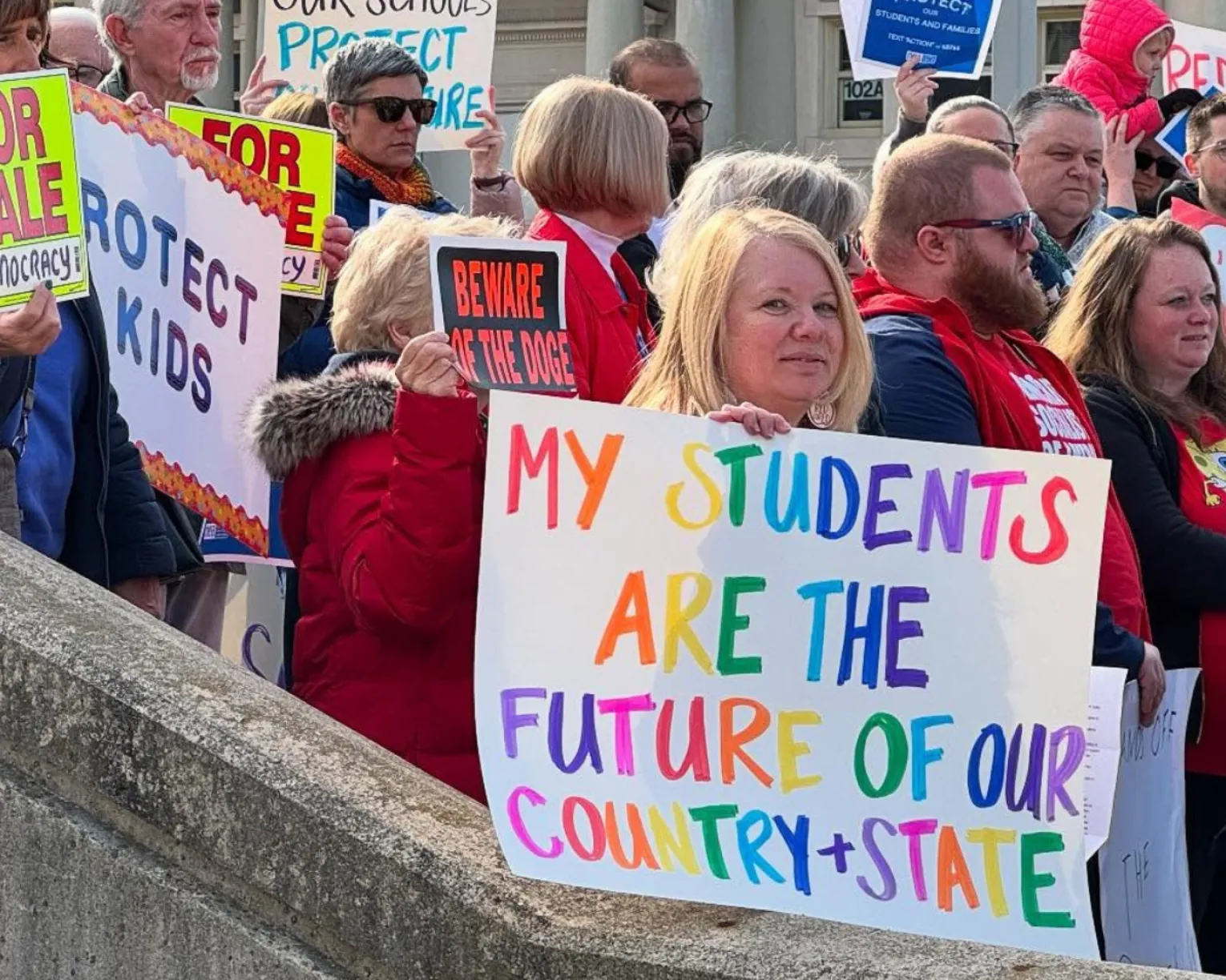At a school walk-in on March 19, 2025, an educator holds a colorful sign that says "My students are the future of our country + state."