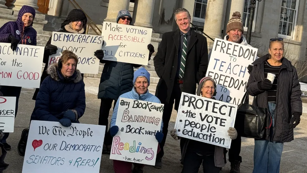 A group of individuals hold protest signs outside the New Hampshire State House